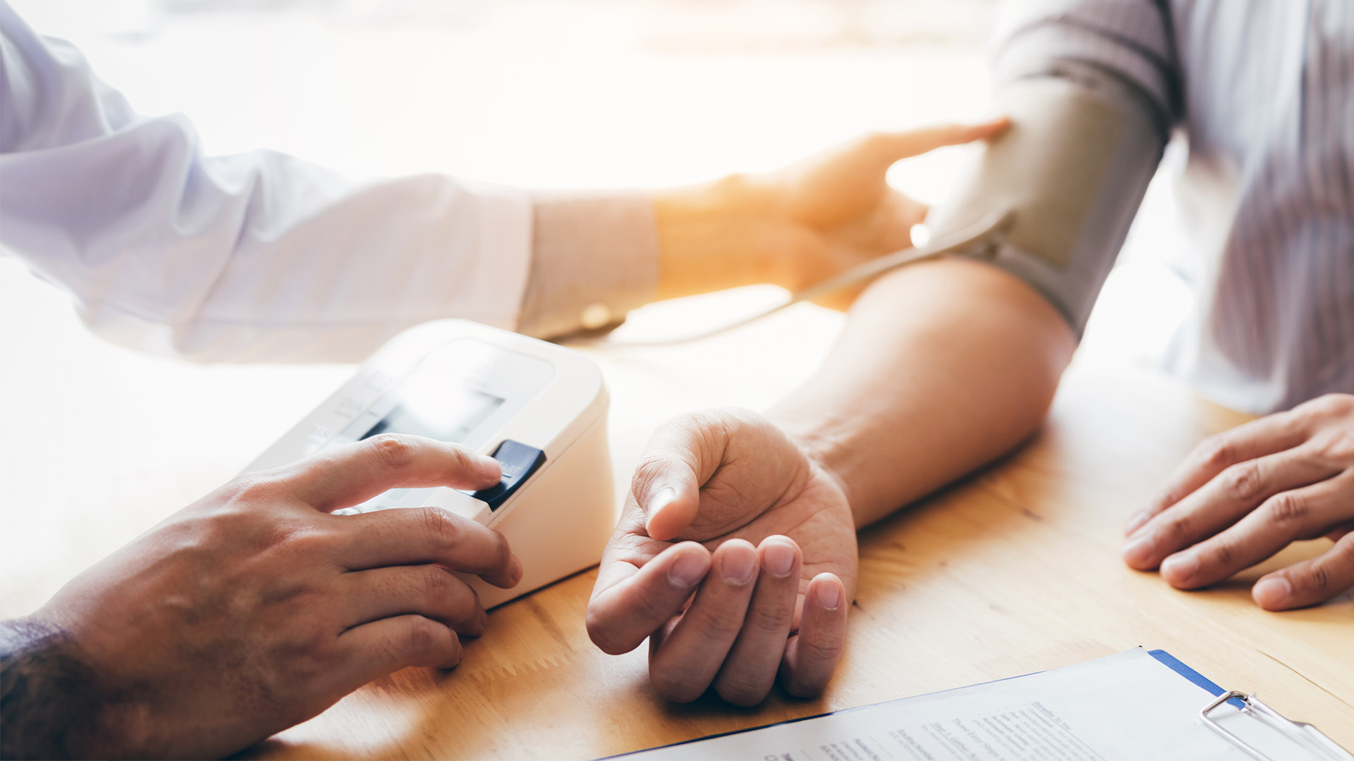 The image shows three individuals in a medical setting a person with a blood pressure cuff around their arm being measured by another individual, while a third person watches, all under the supervision of a healthcare professional.
