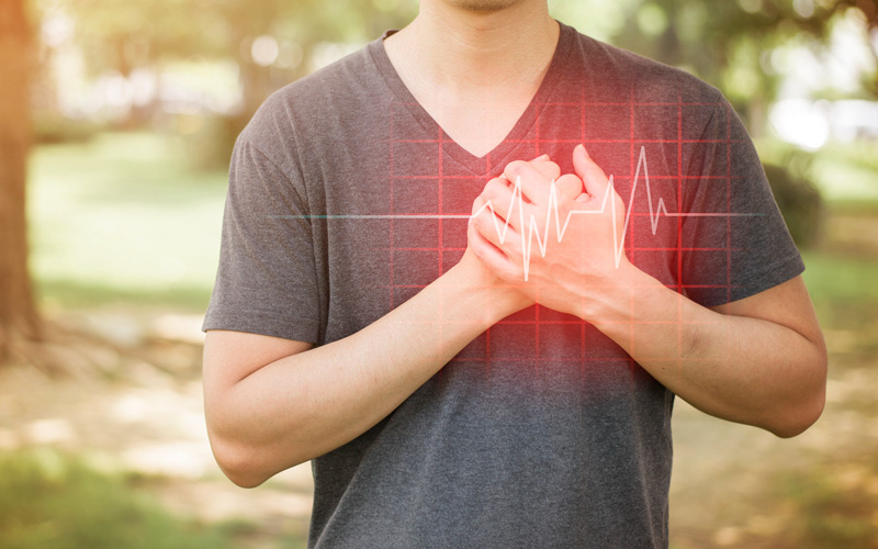 A young man stands outdoors with his hands clasped together in front of him, displaying a heartbeat pattern on his palms through an overlaid digital interface, against a blurred background of trees and grass.